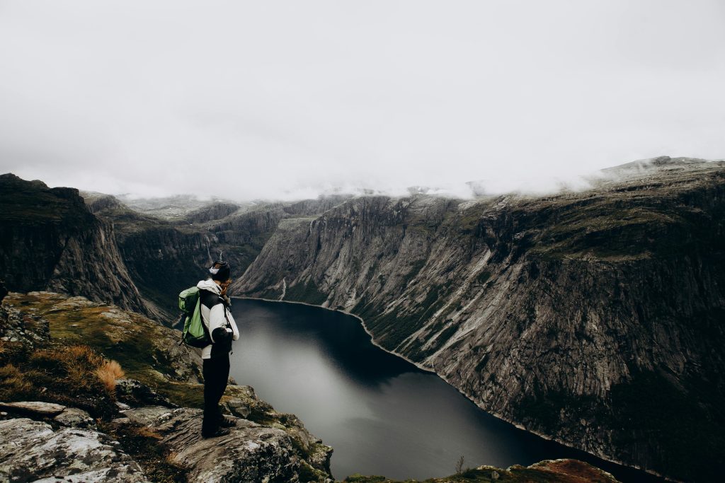 Person standing on a cliff edge overlooking a dramatic fjord landscape in Norway under overcast skies.